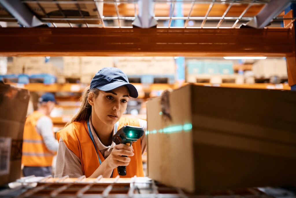 Warehouse worker wearing an orange safety vest and cap scans a barcode on a cardboard box with a handheld scanner while standing between storage racks, with their face blurred for privacy.