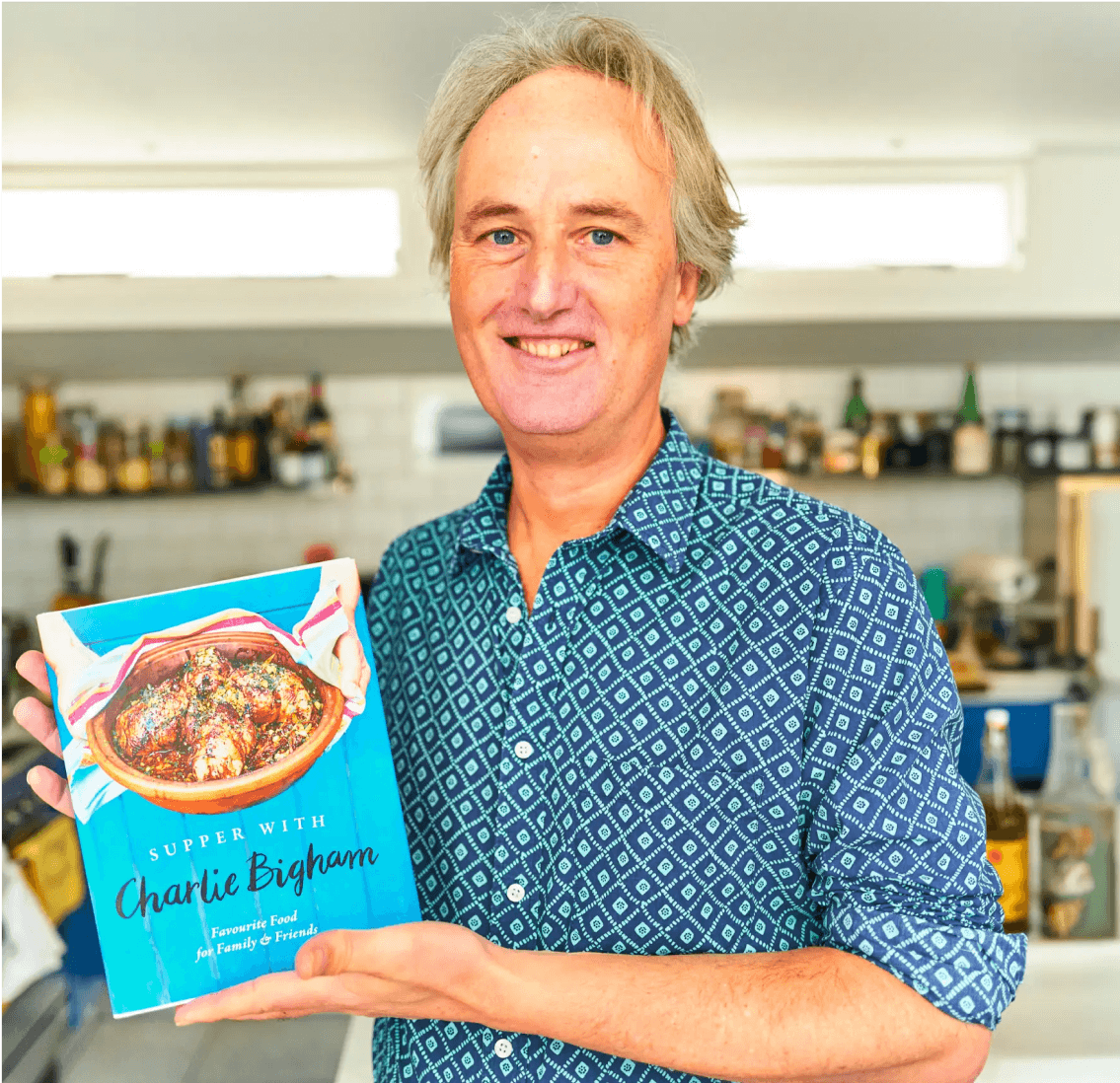 Smiling man in a patterned blue shirt standing in a kitchen and holding a bright blue “Supper with Charlie Bigham” cookbook featuring a roasted dish on the cover, representing the Charlie Bigham’s premium home-cooked food brand.